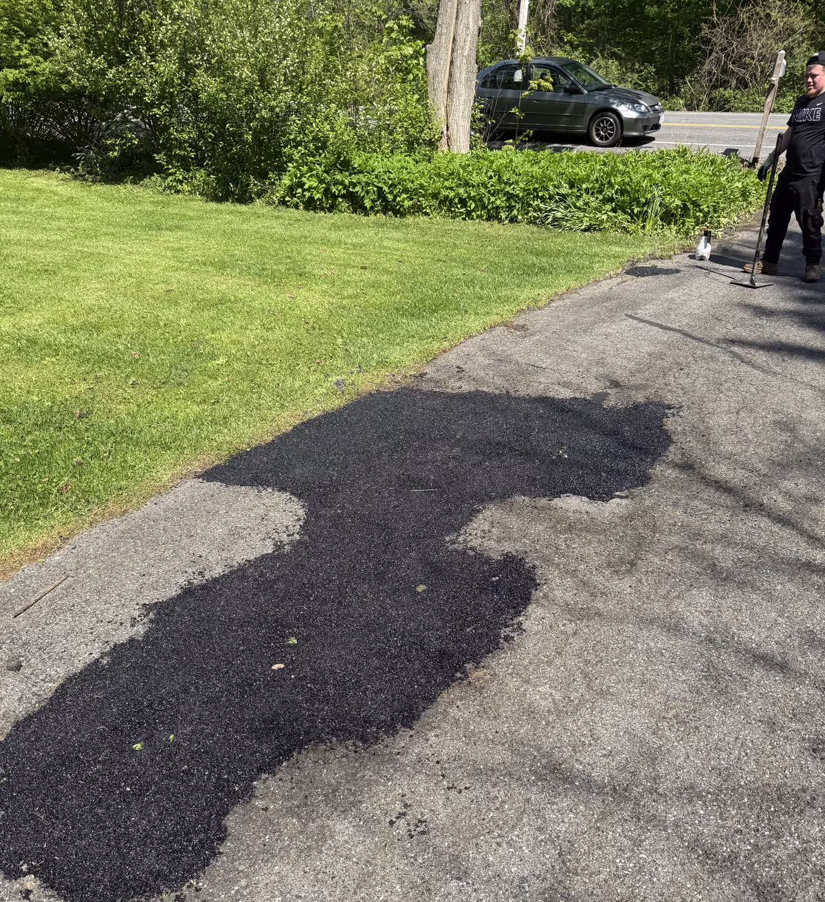 A fresh black asphalt patch laid into a worn gray driveway beside a green lawn