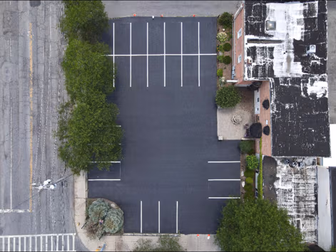 An aerial view of a freshly sealed jet-black commercial parking lot with crisp white lines, tucked between brick buildings and trees