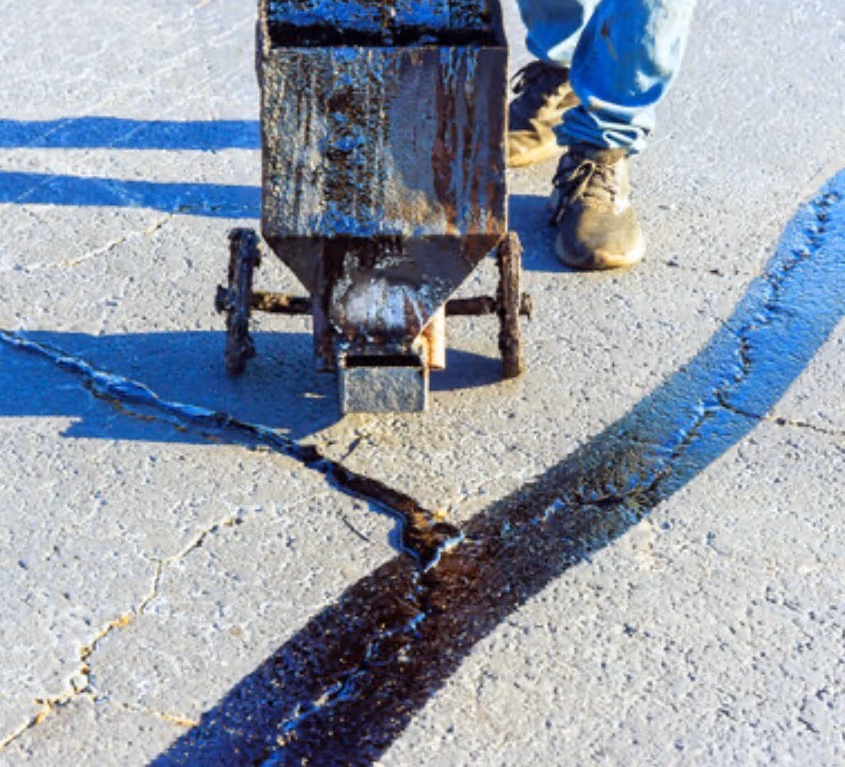 A crack-fill machine laying a flush bead of hot rubber along a long crack in gray asphalt