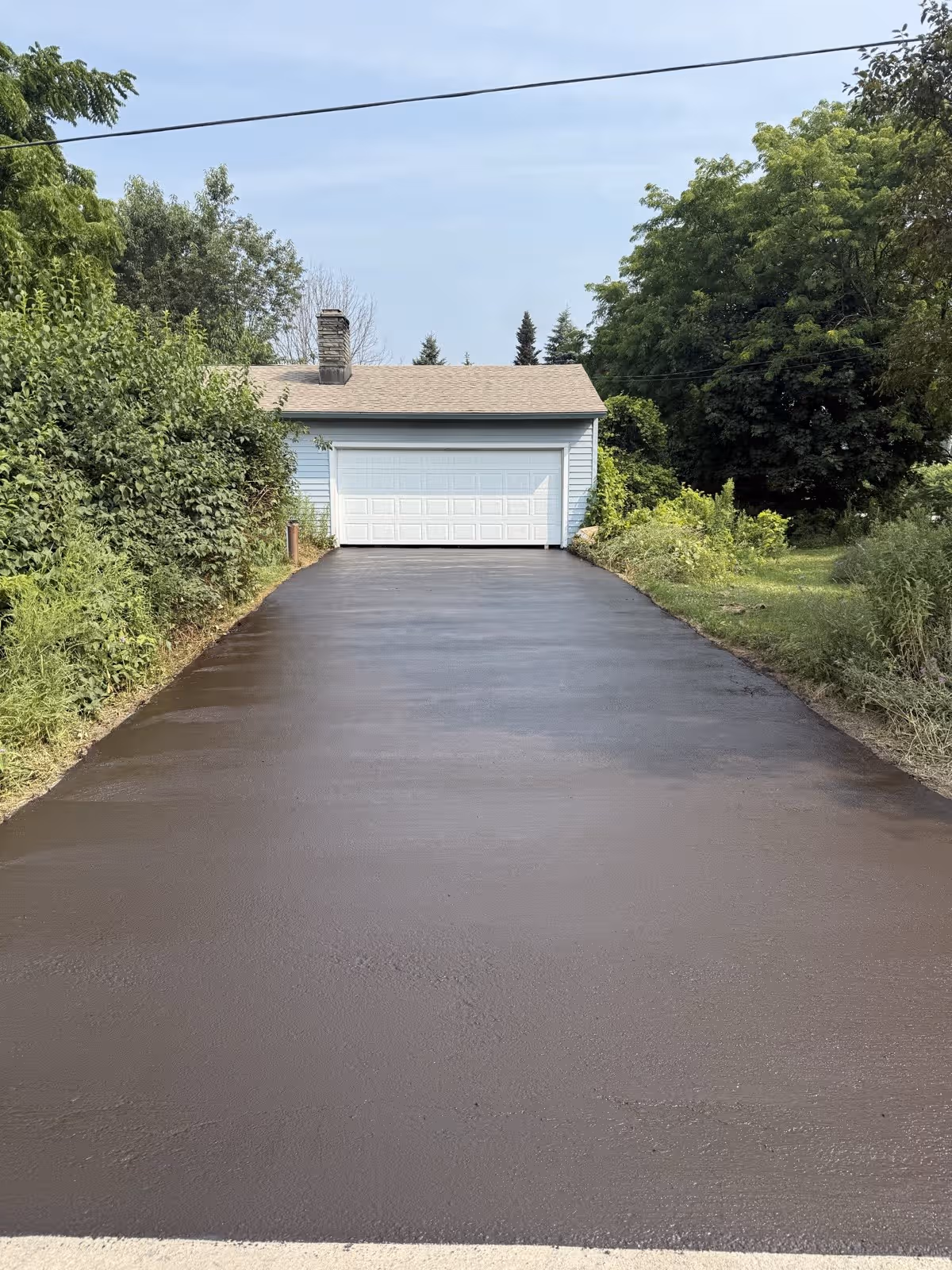 A just-sealed driveway, the dark coat still wet and matte before it cures, leading to a white garage
