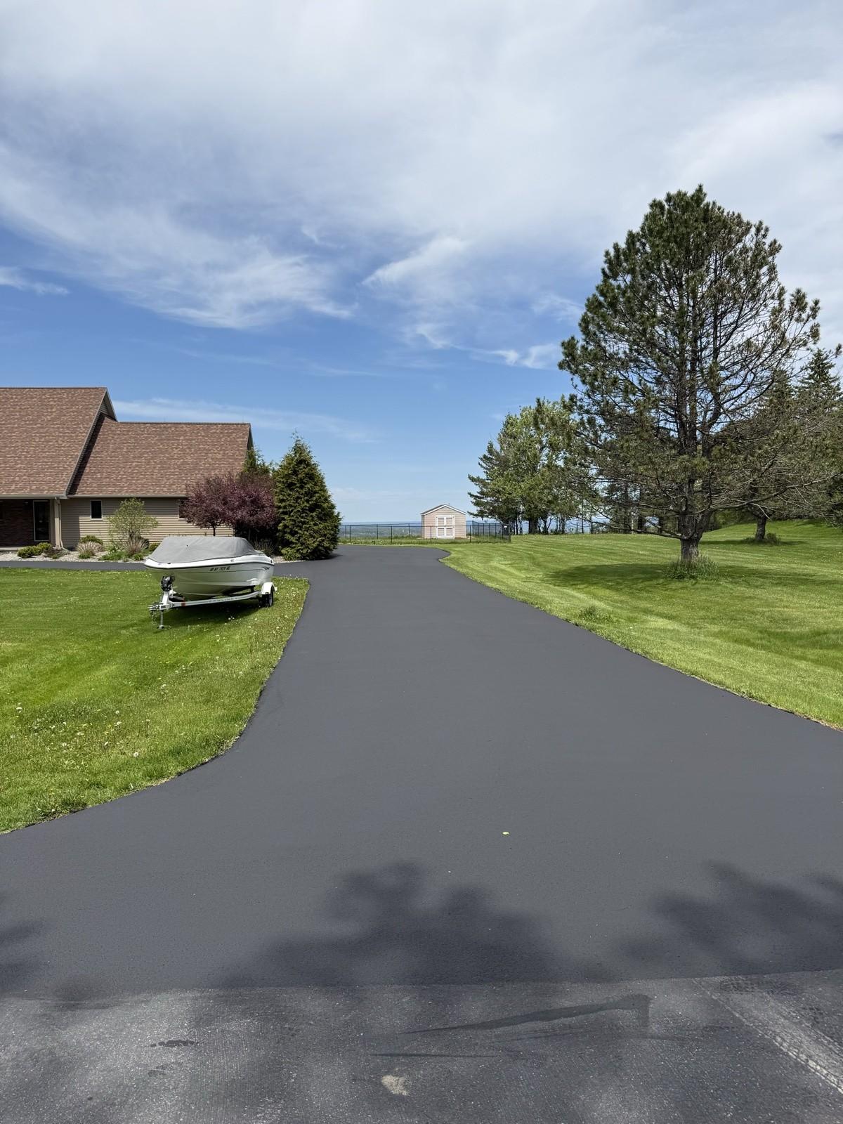 A broad black driveway with room to spare beside a pine tree and a distant lake