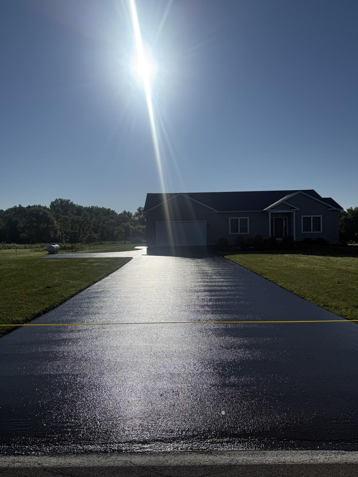 A black sealed driveway beside a gray two-tone house, lit by bright sun and a bold blue sky